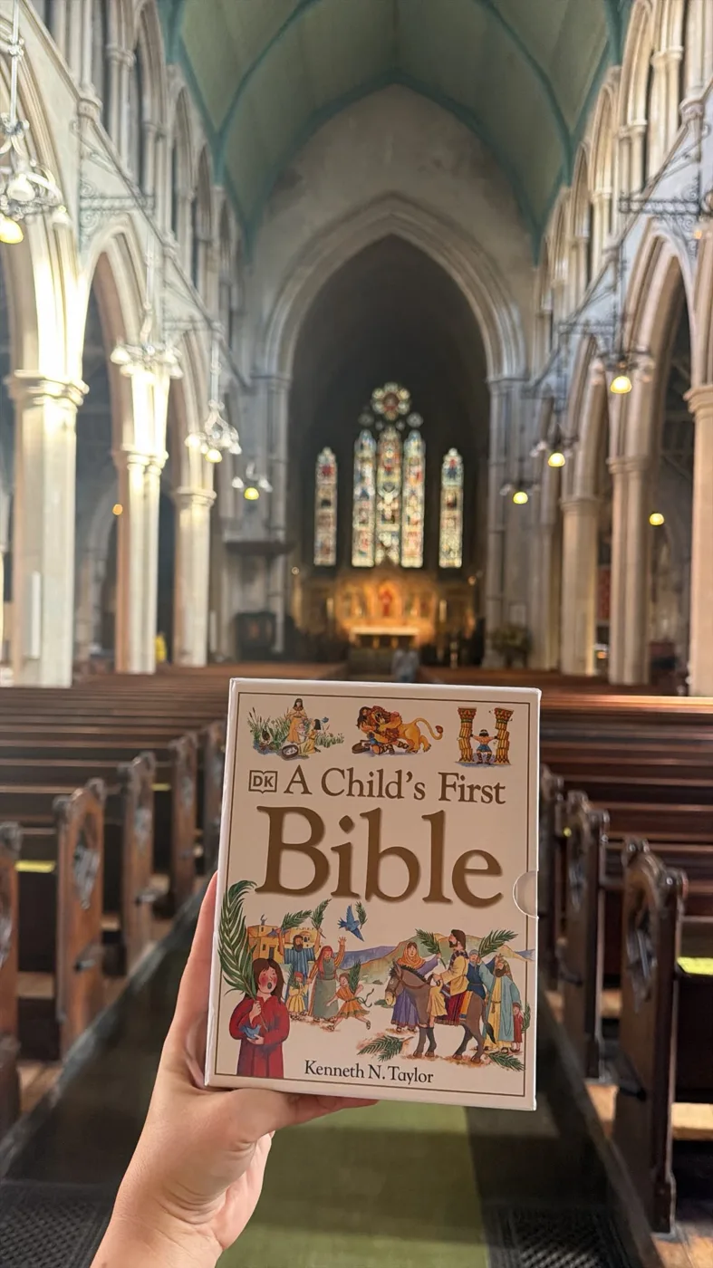 A hand holding up a boxed copy of A Child's First Bible in the nave of St Mary Abbots Church, with rows of pews and the altar window behind