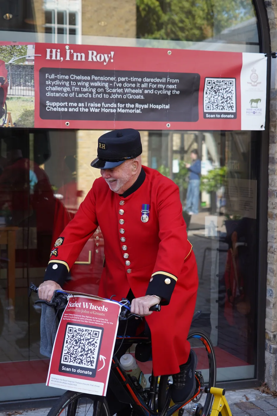 Roy laughs as he pedals the static bike, holding handlebars that carry a Scarlet Wheels sign with a QR code reading Scan to donate