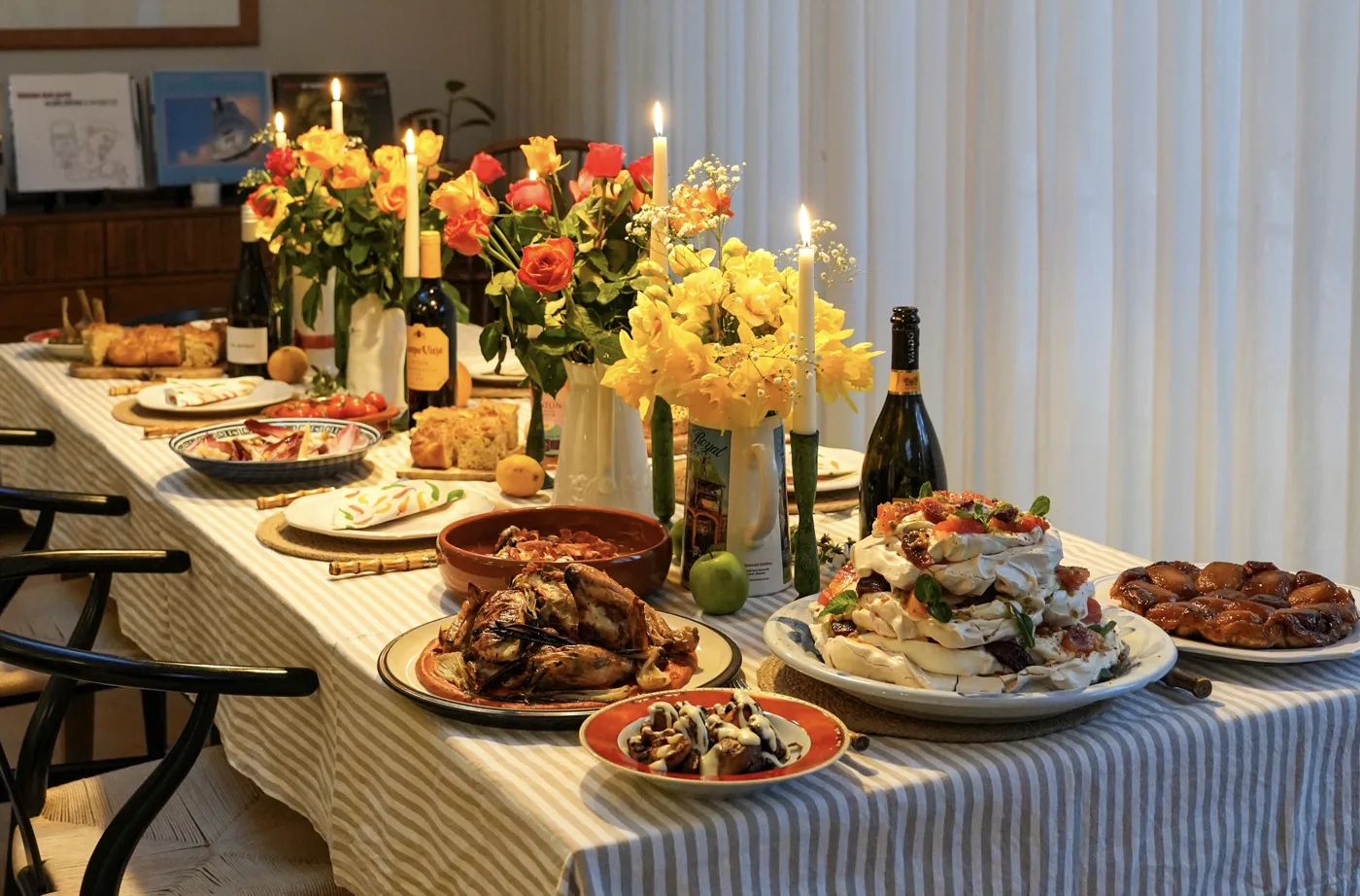 A long dinner table laid with a striped cloth, candles, yellow and orange flowers and sharing plates of food