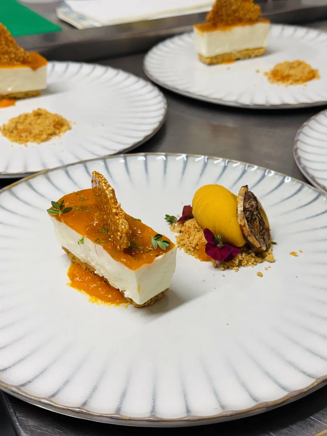 Plated desserts in a row on a steel counter, each a slice with an orange glaze, a crisp shard, crumb and a scoop of orange sorbet