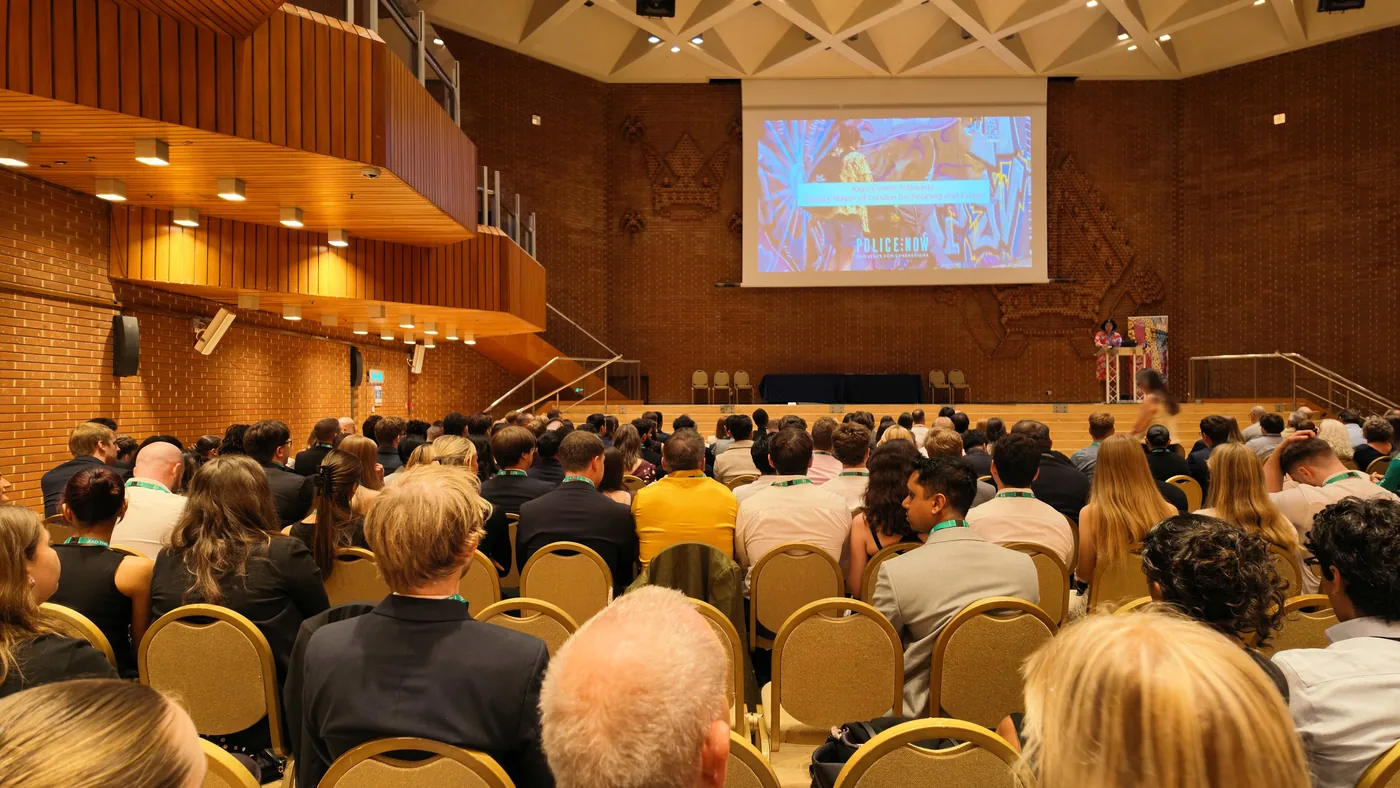 Officers and guests seated in a wood-panelled auditorium facing a stage, with a speaker at a lectern and a Police Now slide on the screen above