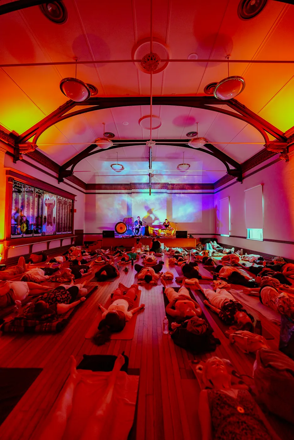 A hall bathed in orange and red light with people lying on mats in front of a performer