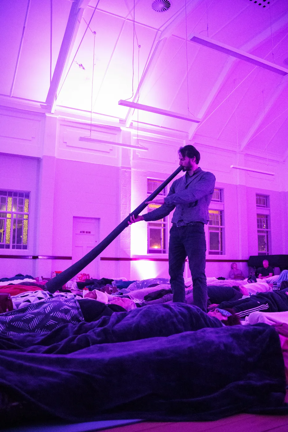 Christian Dimarco standing among people lying under blankets, playing a didjeridu in a hall lit purple