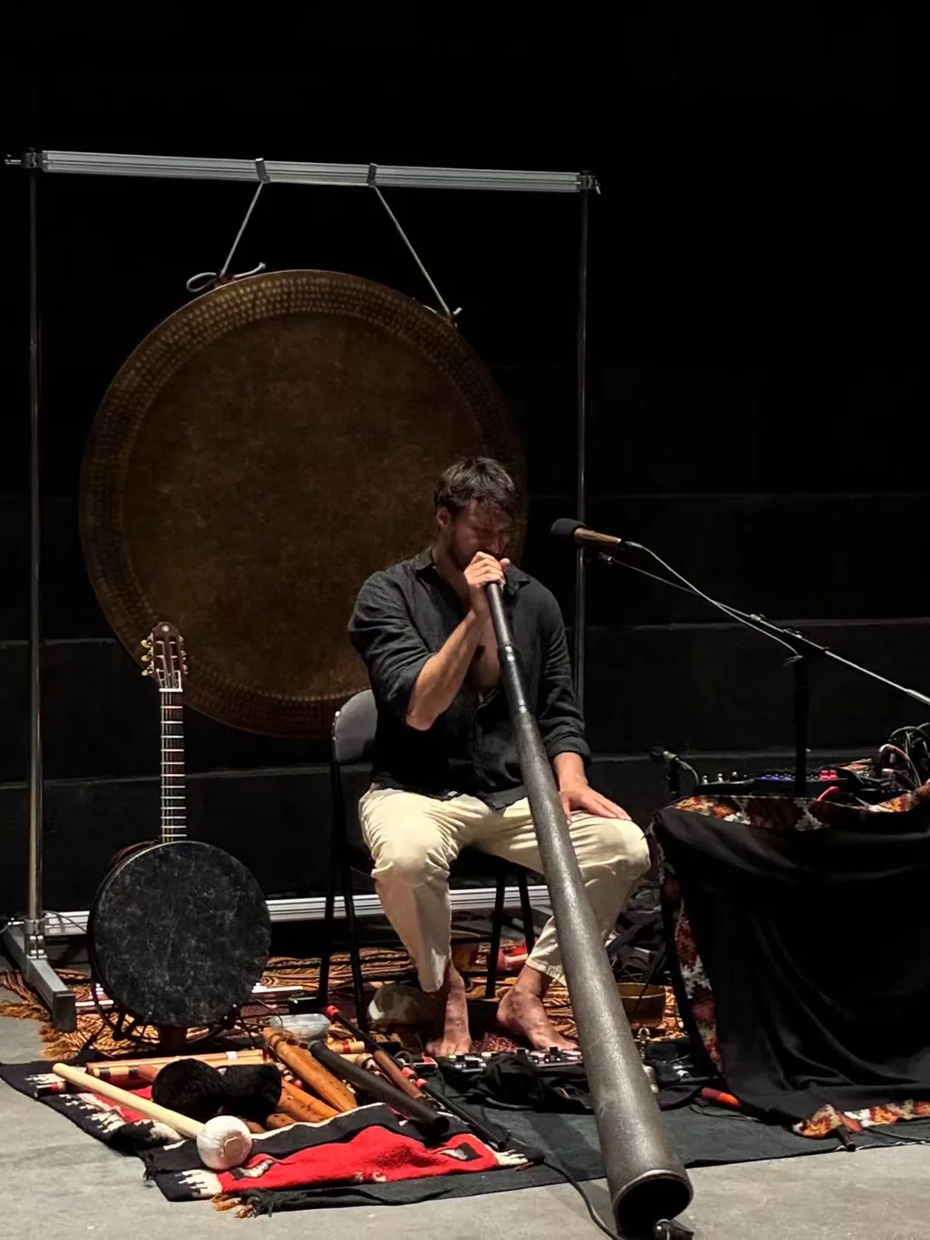 Christian Dimarco seated with a didjeridu in front of a large gong, with flutes, drums and a stringed instrument laid out around him