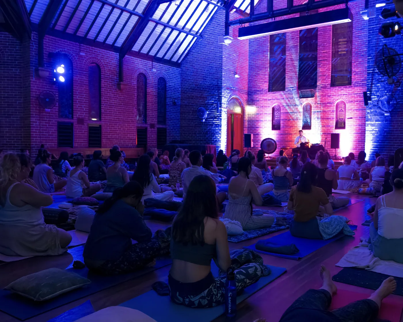 A hall lit in blue and pink with rows of people sitting and lying on mats, facing a small stage
