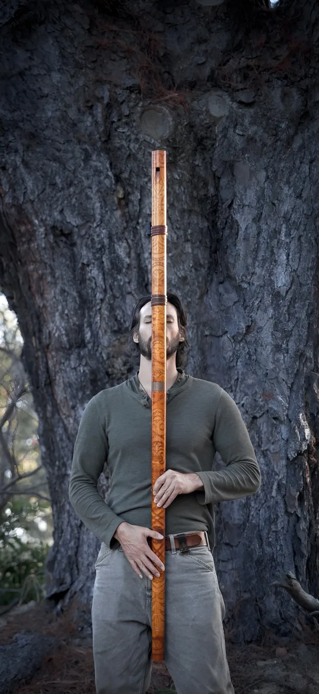 Christian Dimarco standing in front of a tree holding a long wooden flute upright