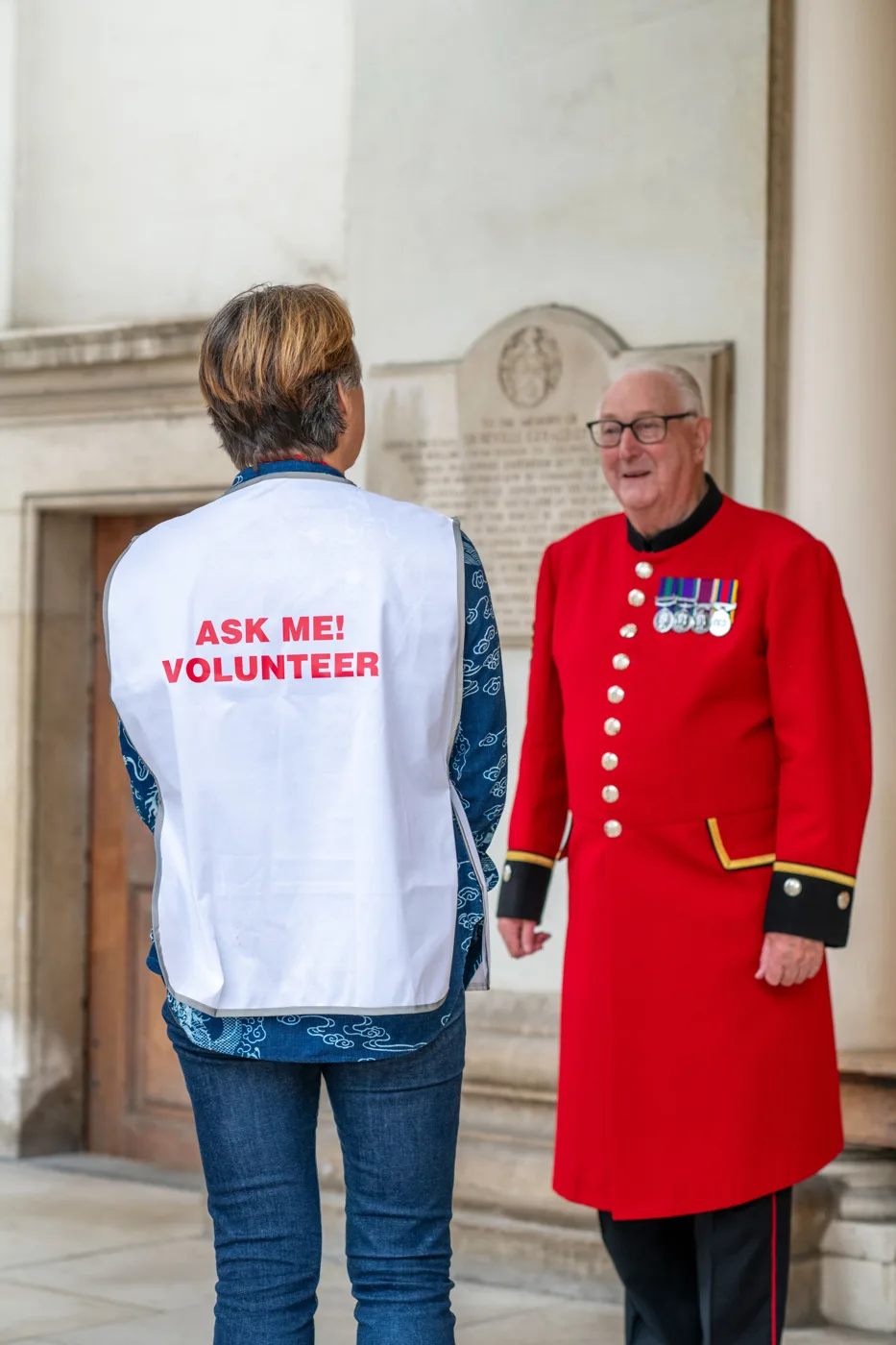 A volunteer in a white tabard reading Ask me! Volunteer, seen from behind, talks with a Chelsea Pensioner in a scarlet coat and medals