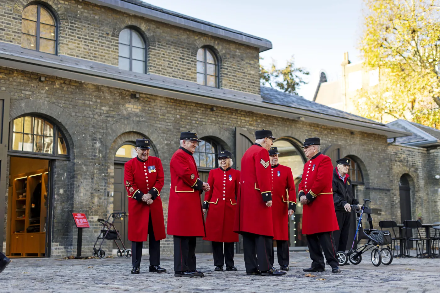 Eight Chelsea Pensioners, most in scarlet coats and black caps, talk in a cobbled yard in front of an arched brick building, with two walking frames beside them
