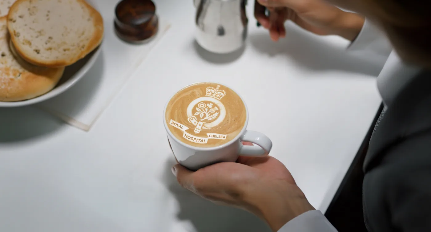 A hand holds a white cup of coffee with the Royal Hospital Chelsea crest drawn in the foam, beside a plate of toasted muffins