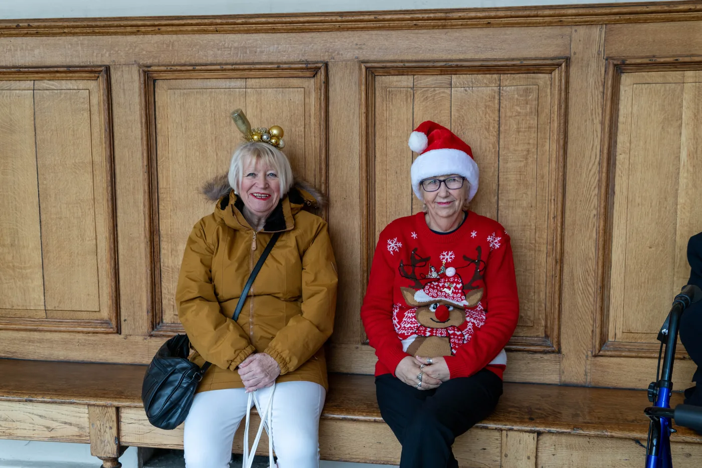 Two women sit on a wooden bench against wood panelling, one in a mustard coat wearing a sparkly reindeer-antler headband, the other in a red Christmas jumper and Santa hat