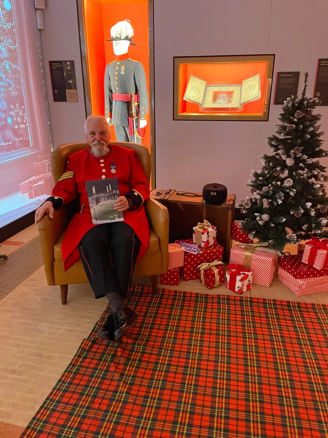 A Chelsea Pensioner in a scarlet coat sits in a yellow armchair reading a book, beside a Christmas tree with wrapped presents on a red tartan rug
