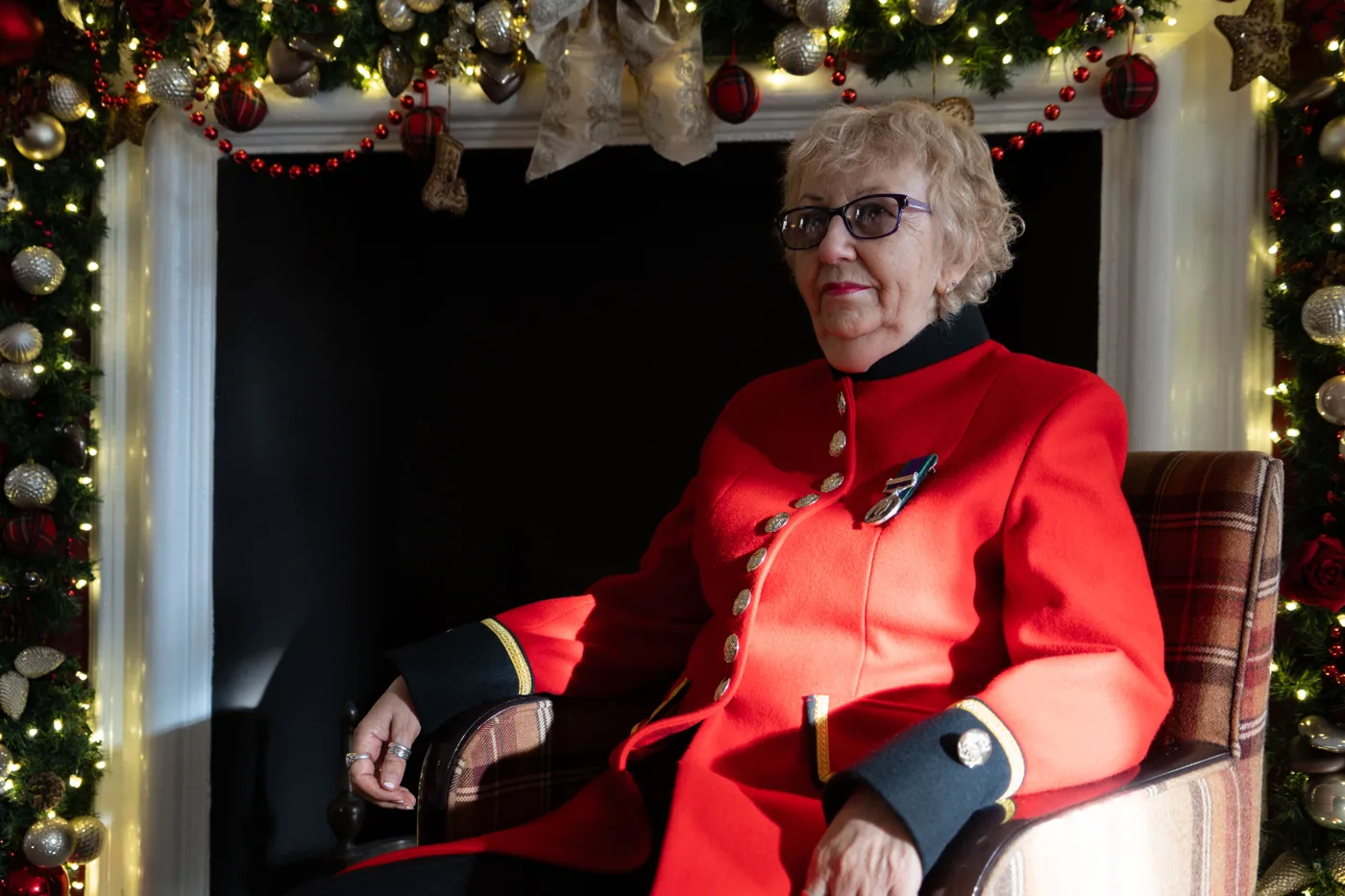 A woman Chelsea Pensioner in a scarlet coat and glasses sits in an armchair framed by a fireplace hung with a garland of baubles and lights