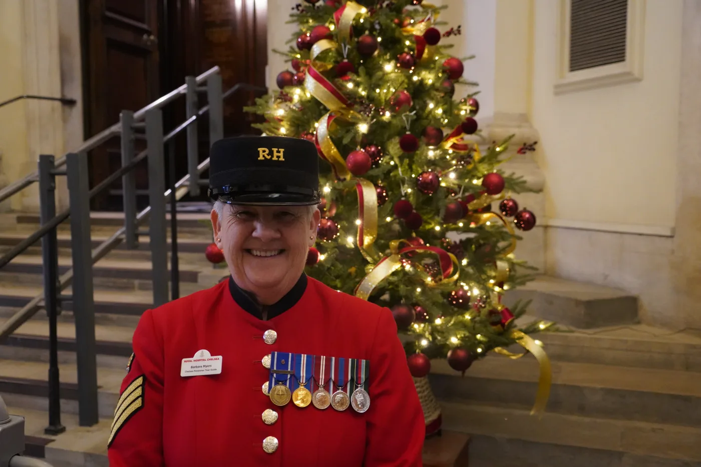 A smiling Chelsea Pensioner in a scarlet coat with a row of medals and a black RH cap stands on stone steps beside a decorated Christmas tree