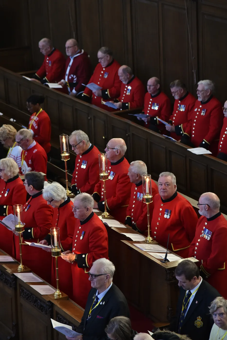 Rows of Chelsea Pensioners in scarlet coats seated along wooden benches in a panelled hall