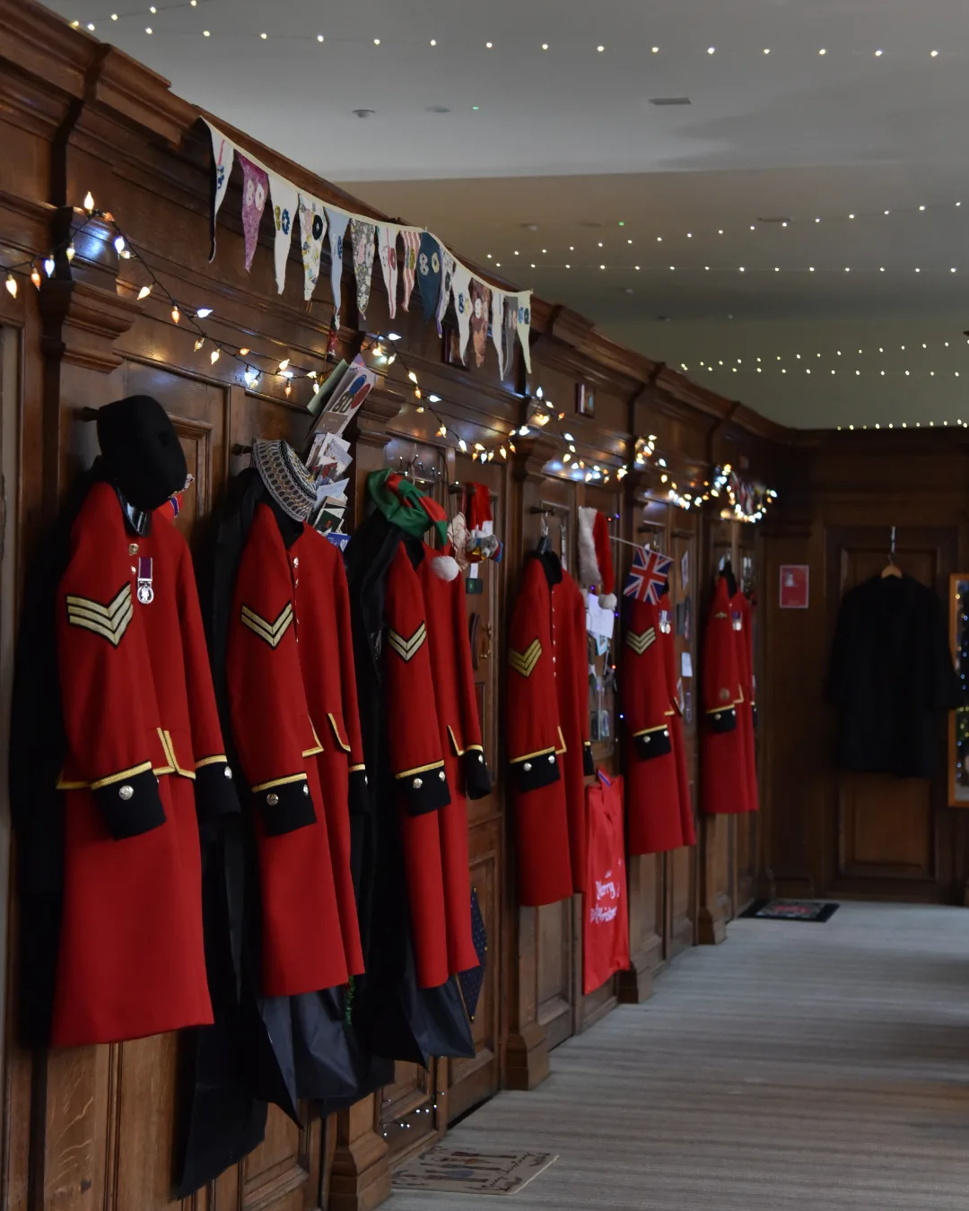 A row of scarlet Chelsea Pensioner coats and black caps hanging on pegs along a wood-panelled corridor strung with bunting and fairy lights