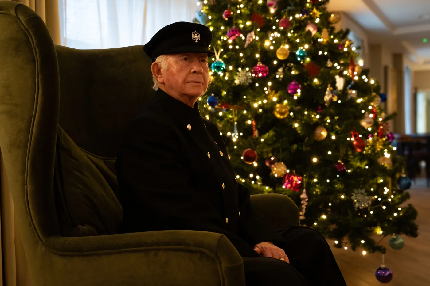 A Chelsea Pensioner in a dark blue uniform and cap sits in a green armchair beside a tall Christmas tree hung with coloured baubles and lights