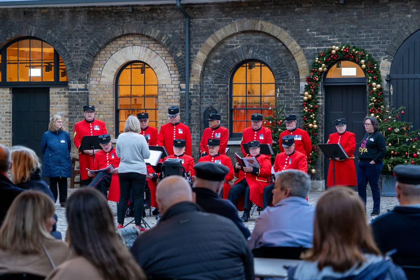 Chelsea Pensioners in scarlet coats and black caps sit and stand with songbooks and music stands in a brick courtyard, singing to a seated audience, with a conductor in front of them and a wreathed arch and Christmas tree to the right