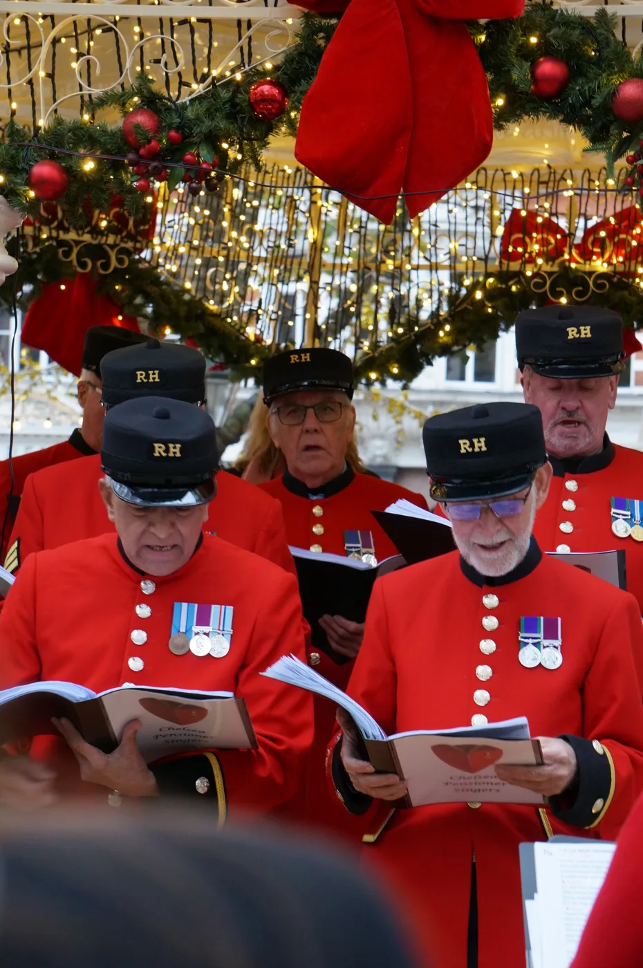 A close group of Chelsea Pensioners in scarlet coats and black caps reading from songbooks under a garland of red bows and lights