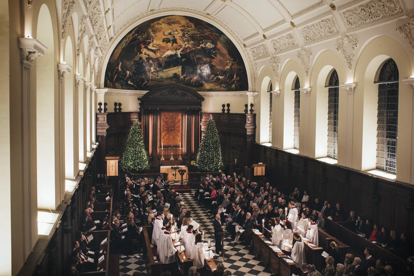 View from the gallery of the Wren Chapel during a carol service, with a choir in white surplices in the centre, a full congregation in the pews, and two lit Christmas trees beneath the painted apse