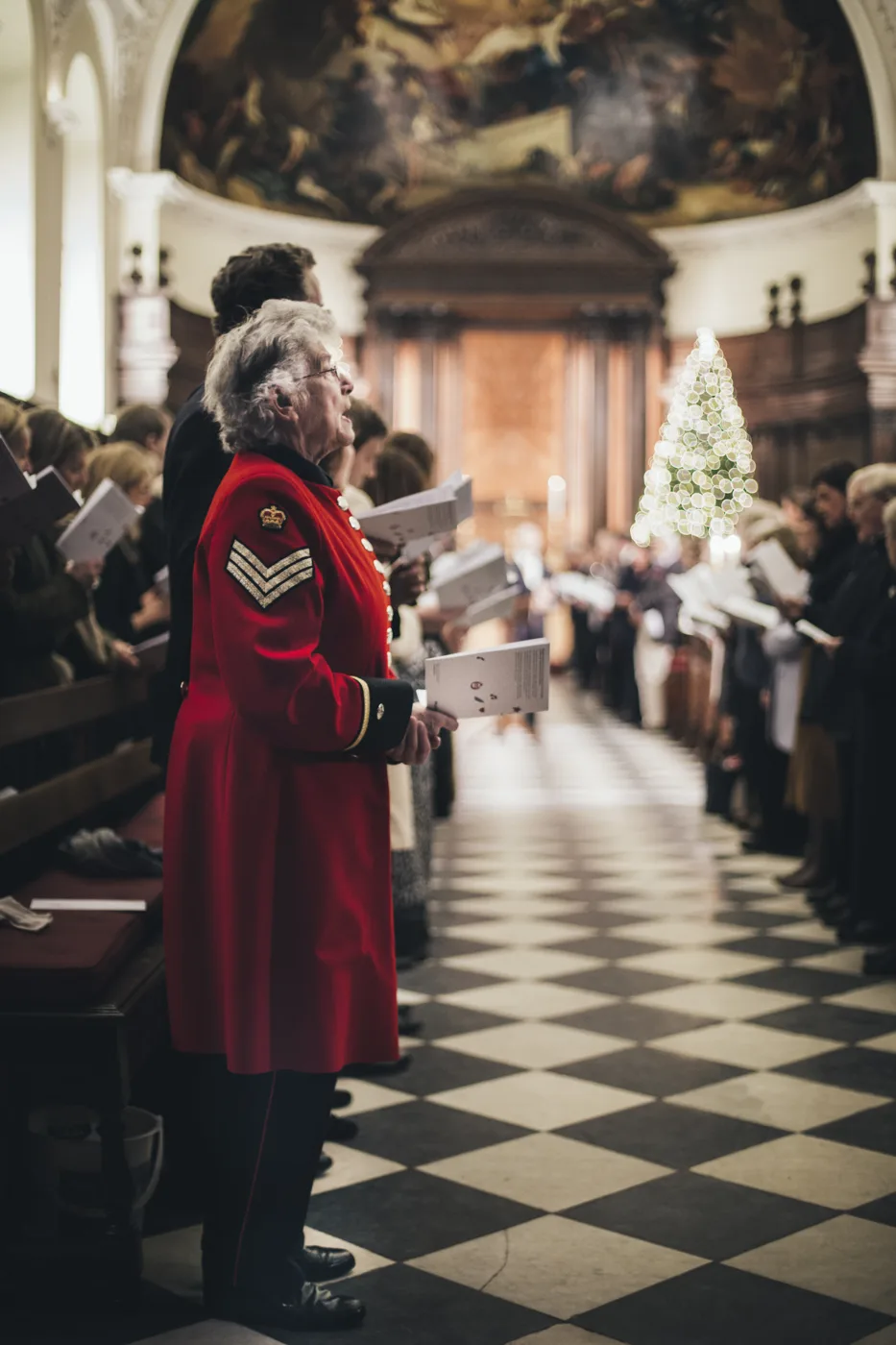 A Chelsea Pensioner in a scarlet coat with medals stands in the chequered aisle of the Wren Chapel holding a service sheet, with the congregation and a Christmas tree behind him