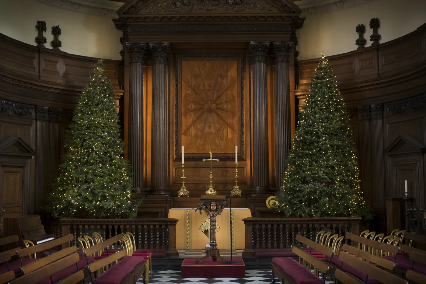 The altar of the Wren Chapel framed by two tall Christmas trees covered in white lights, with candlesticks on the altar and panelled pews either side