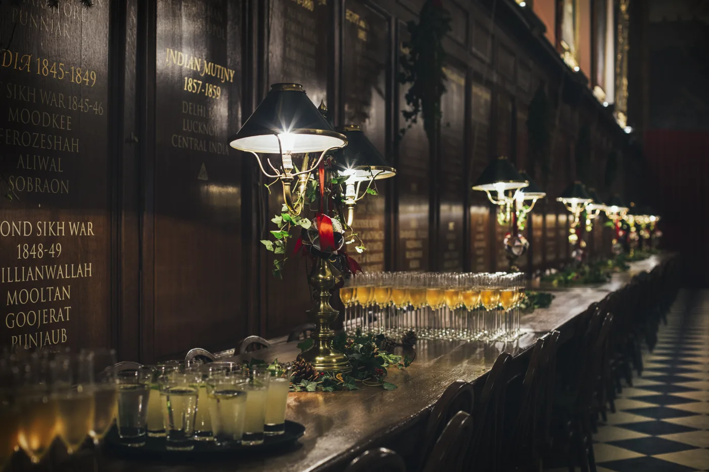 A long dark wooden table in the Great Hall laid with rows of glasses, evergreen garlands and brass lamps with green shades, with wooden panels of battle honours on the wall