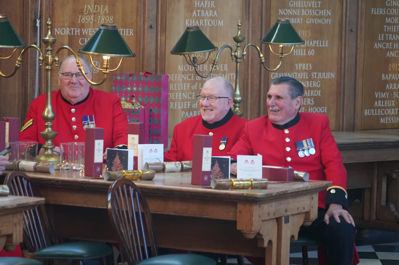 Three smiling Chelsea Pensioners in scarlet coats with medals sit at a long wooden table laid with Christmas crackers, cards and gift boxes, beneath brass lamps and wooden panels carved with battle honours