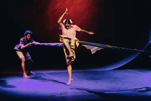 A dancer leaps on a dark stage while a second performer pulls a long sheet of fabric behind her