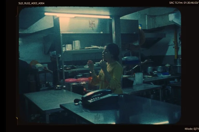 A film still of a woman in a yellow top standing in a steel commercial kitchen