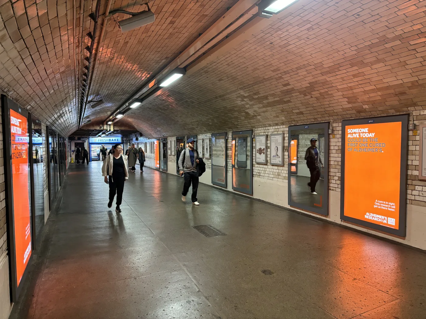 Mirrors and orange Alzheimer's Research UK panels along both walls of the tiled subway at South Kensington station