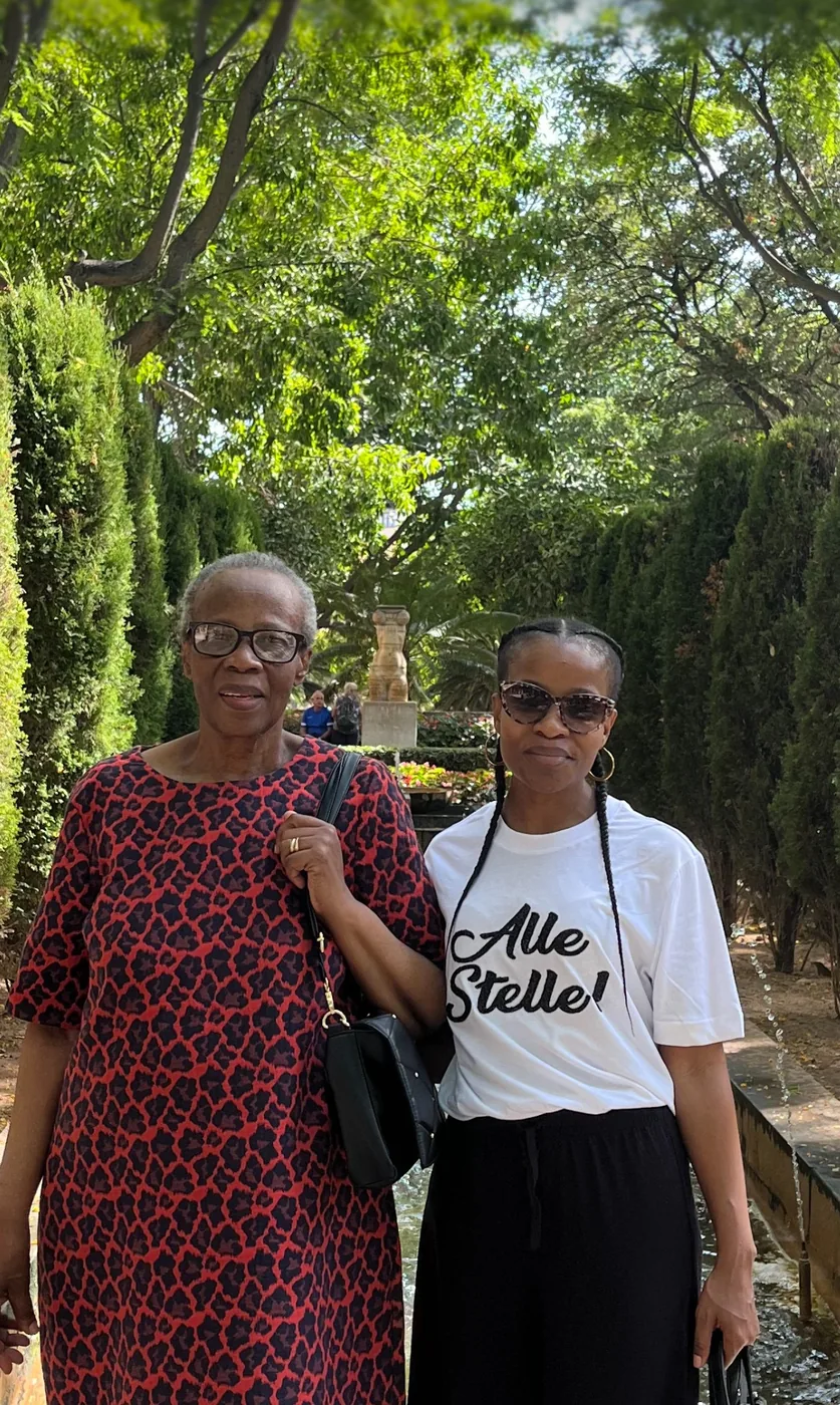 Edith and Dineo standing together on a garden path lined with trees and clipped conifers